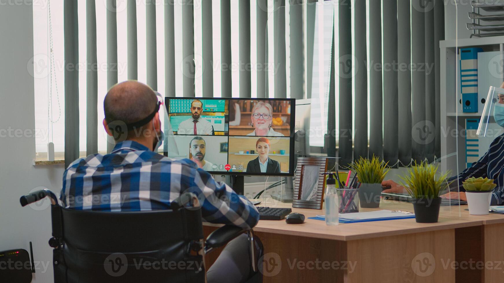 Paralysed manager in wheelchair talking with mask and visor during videocall having online conference in business office. Immobilized freelancer working in financial company respecting social distance photo