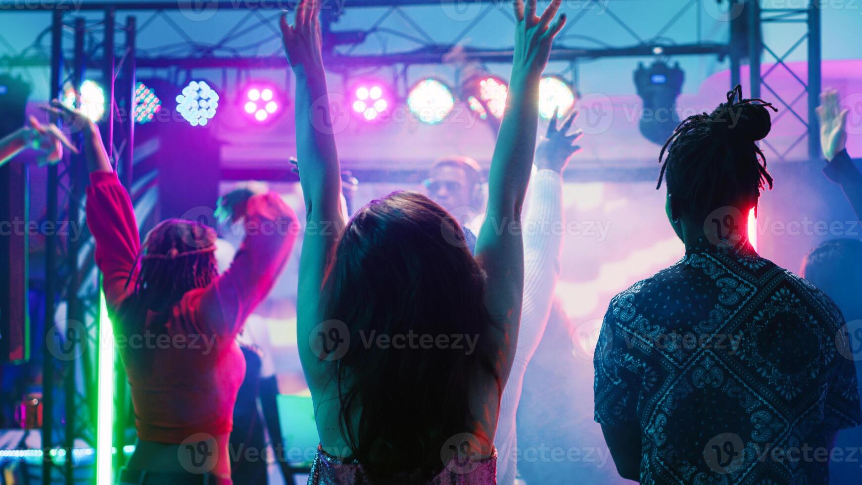 Diverse people partying at nightclub, enjoying live music performance from DJ station at discotheque stage. Group of friends dancing together on dance floor with spotlights. Tripod shot. photo