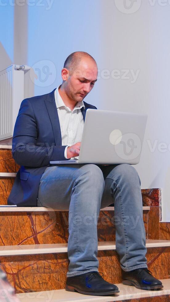 Overworked tired businessman at workplace working on deadline for job project using notebook sitting on stairwell. Confident corporate entrepreneur using laptop doing overtime in business building. photo