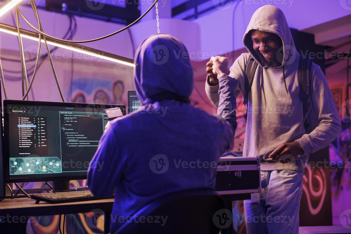Cheerful african american hacker giving fist bump to crime partner while working together in abandoned warehouse. Smiling men in hoods sharing friendly gesture after successful database breaking photo