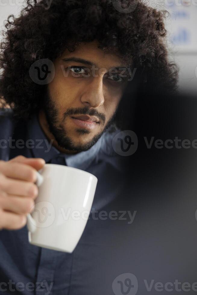 Start up company business employee drinking cofee and doing product management on laptop. Young arab entrepreneur holding tea cup and preparing marketing campaign on computer photo