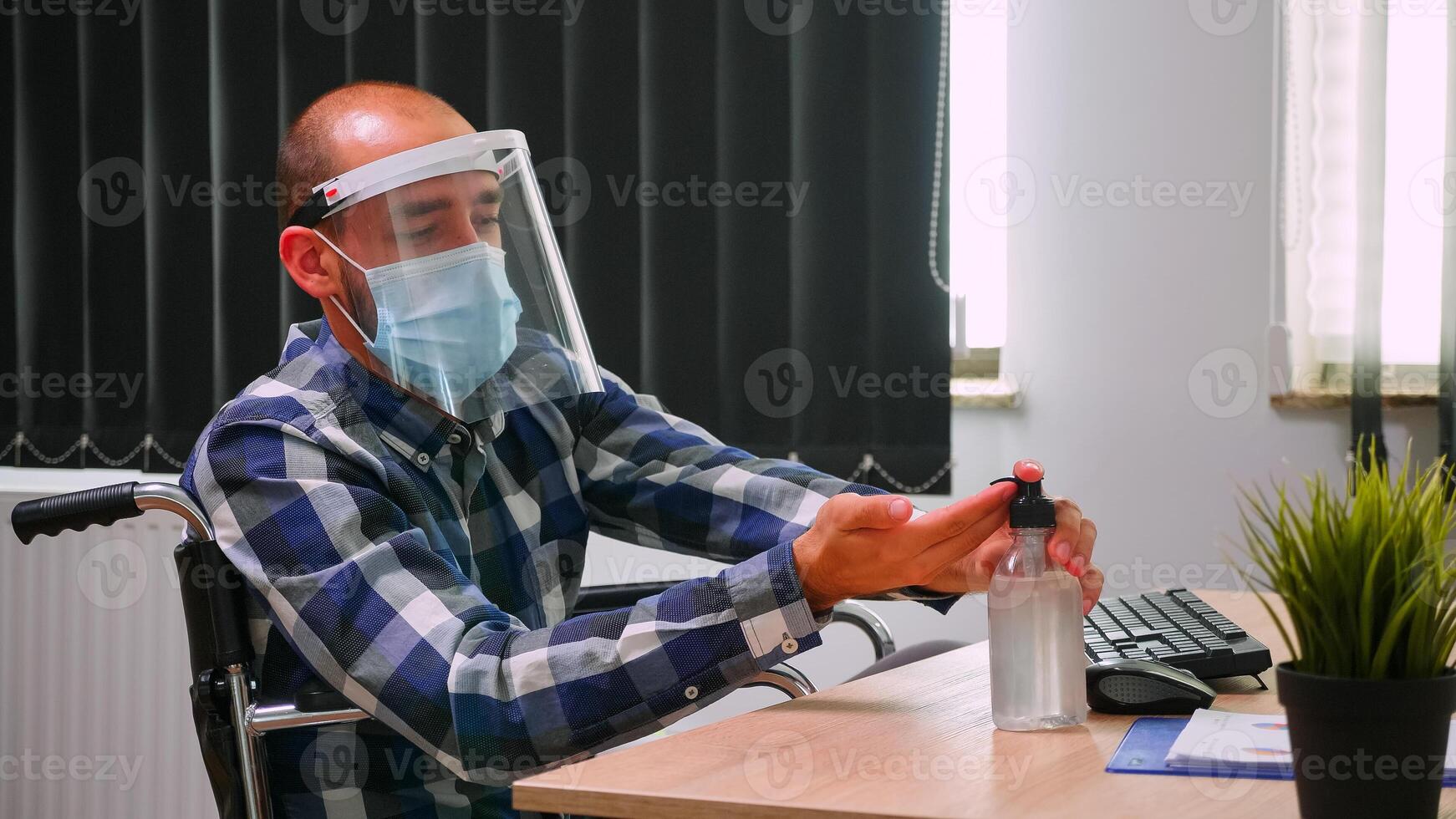 Disabled entrepreneur sitting in wheelchair with protection mask cleaning hands before typing on computer in business modern office. Handicapped freelancer with visor respecting social distance photo