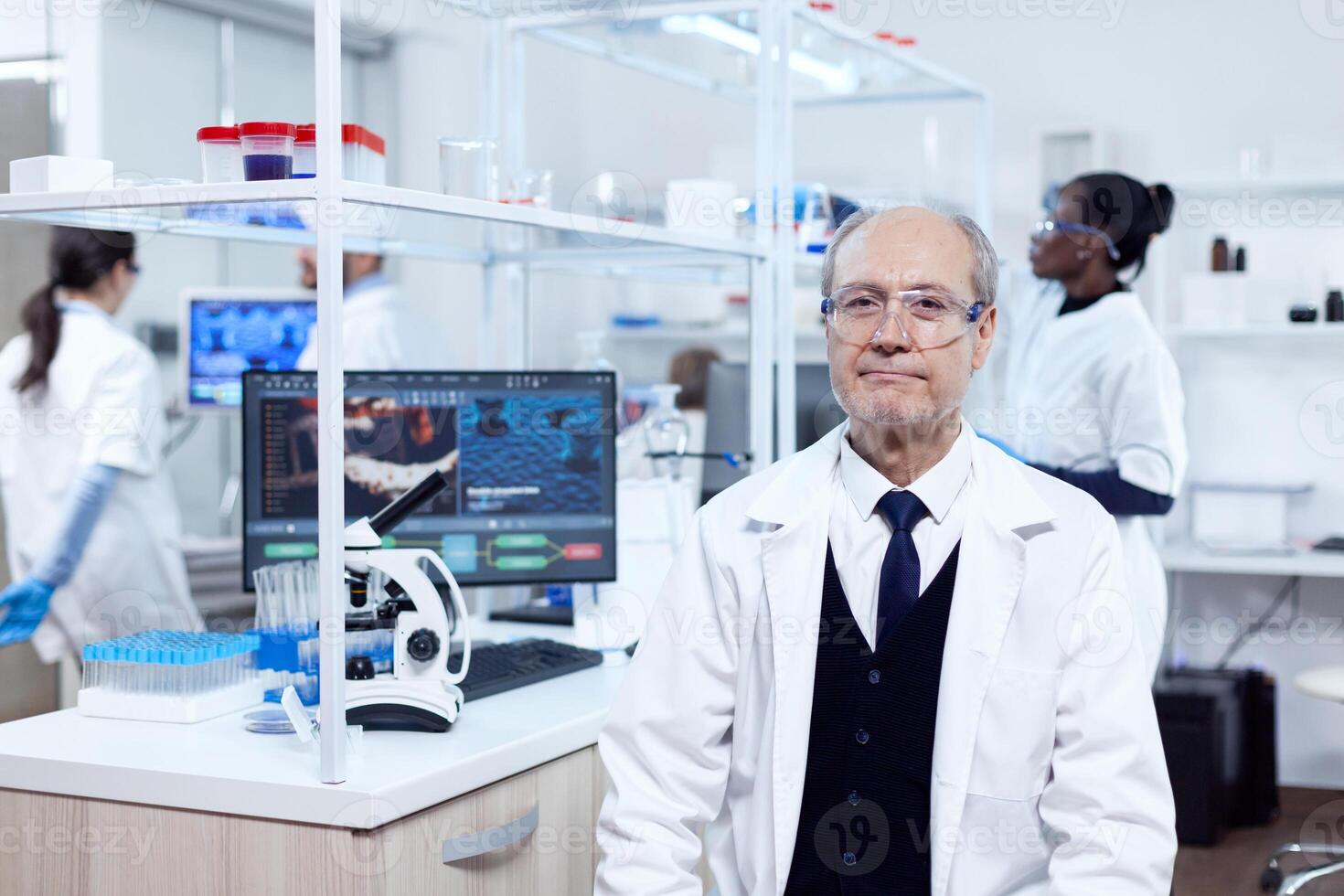 Senior man with virus on computer display looking at camera with protective glasses. Elderly scientist wearing lab coat working to develop a new medical vacine with african assistant in the background. photo