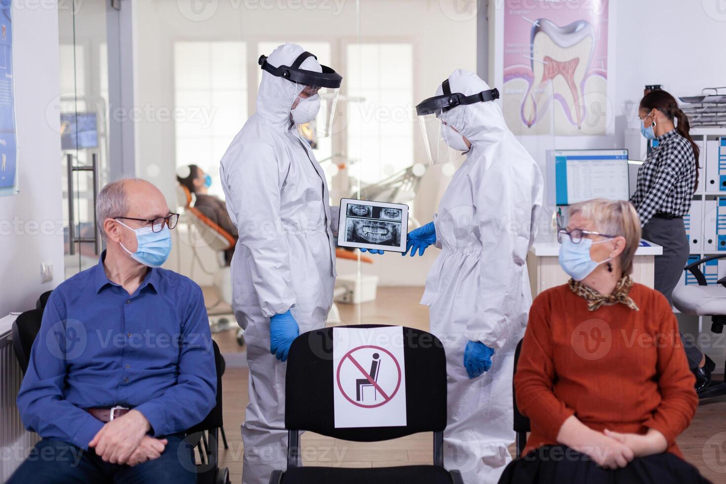 Man discussing with nurse in dental reception wearing protection suit against coronavirus, elderly patients waiting in reception keeping distance. Concept of new normal dentist visit in outbreak. photo
