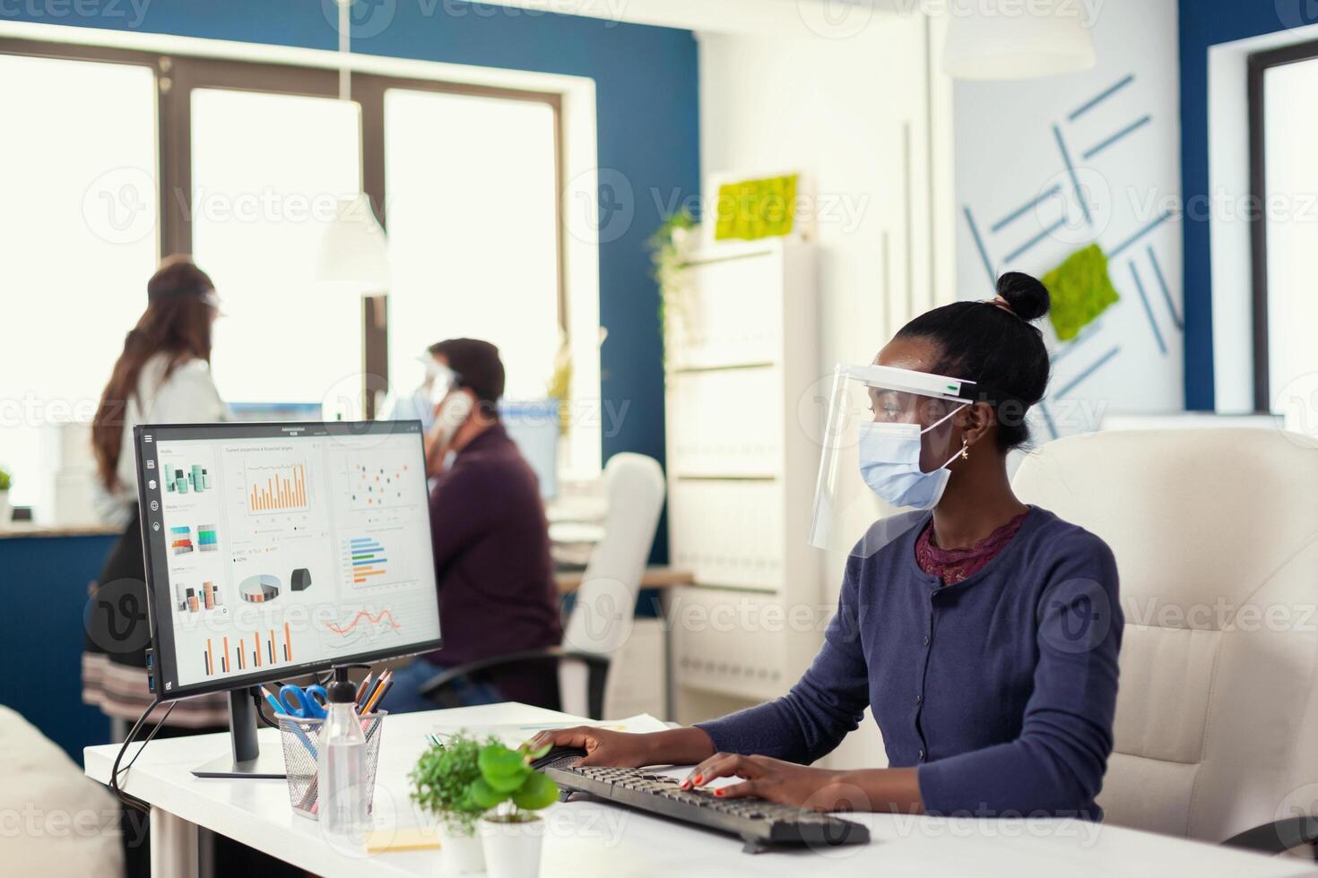 African woman at workplace working on computer looking at graph wearing face mask against covid19. Multiethnic team in new normal business financial office checking reports, analysing datas looking at desktop. photo