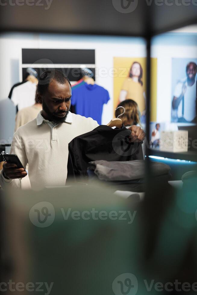 African american man shopping for shirt and checking apparel in stock using smartphone online app. Clothing store customer holding outfit on rack and scrolling website on mobile phone photo