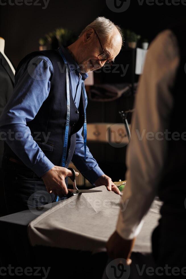 Smiling elderly master suitmaker and skilled tailor apprentice cutting fabric sheet for upcoming stylish fashion collection. Couturiers in atelier manufacturing refined sartorial garments photo