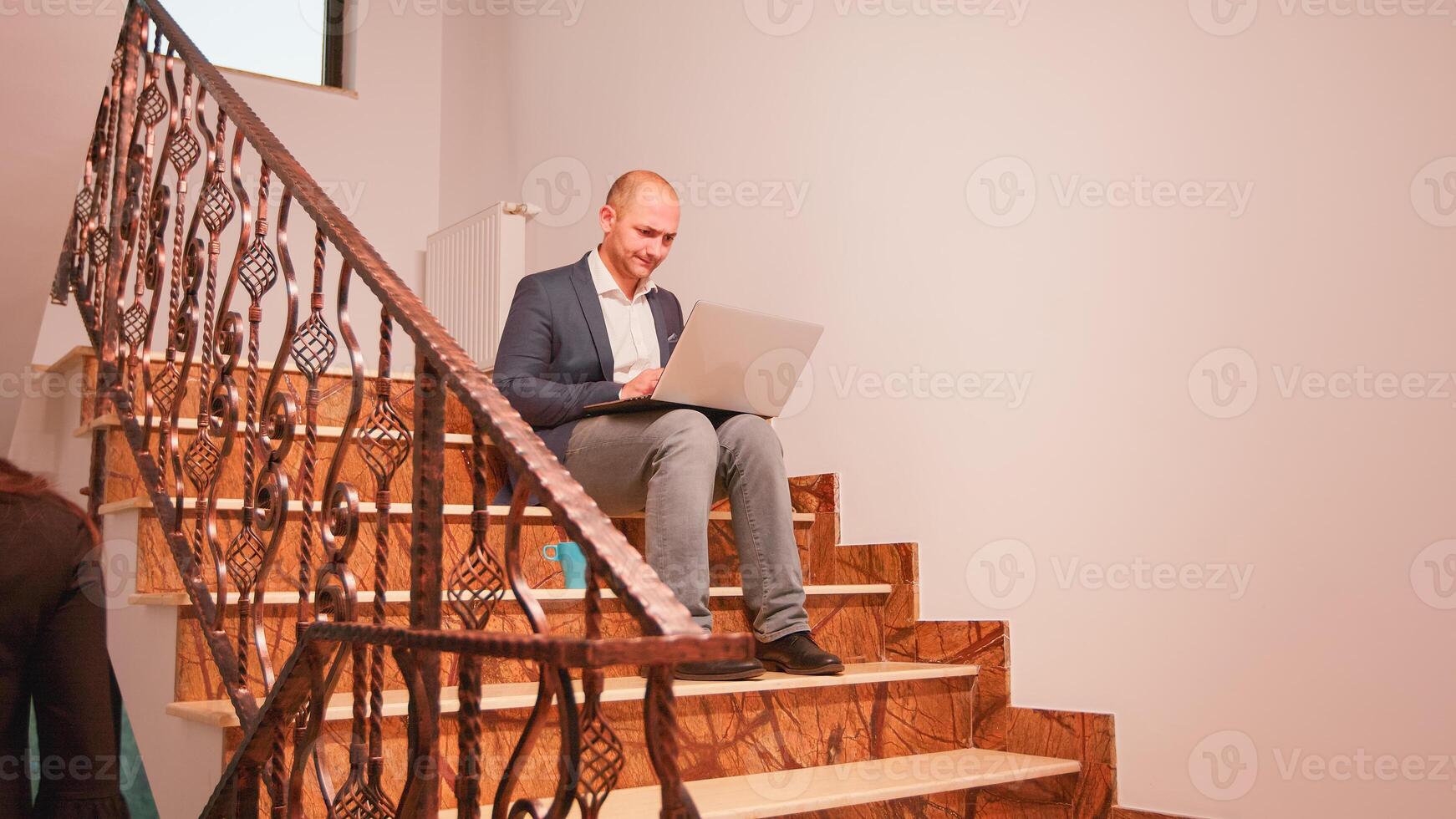 Exhausted businessman using laptop working alone on difficult deadline sitting on stair overtime. Executive manager doing overtime at corporate job on staircase while colleagues leaving office building photo