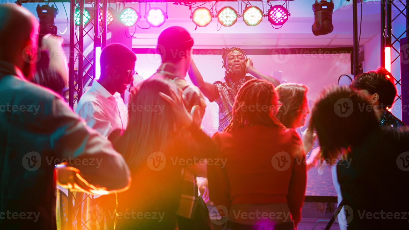 Multiethnic group of people partying on dance floor, enjoying electronic funky music from DJ mixing station. Cheerful adults having fun at nightclub, dancing together at celebration. Handheld shot. photo