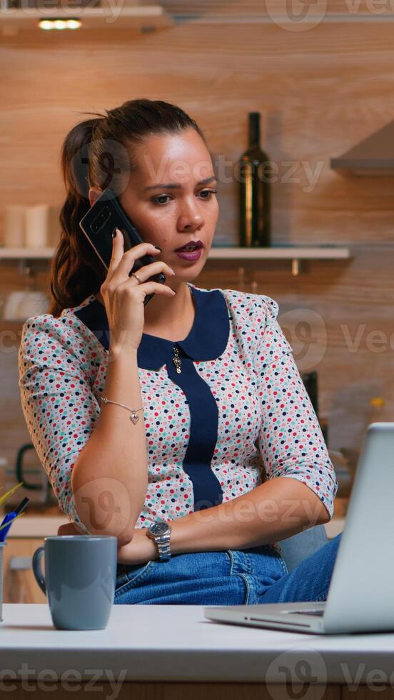 Remote employee speaking at phone while working at laptop late at night. Busy focused freelancer using modern technology network wireless doing overtime for job reading writing, searching taking break photo