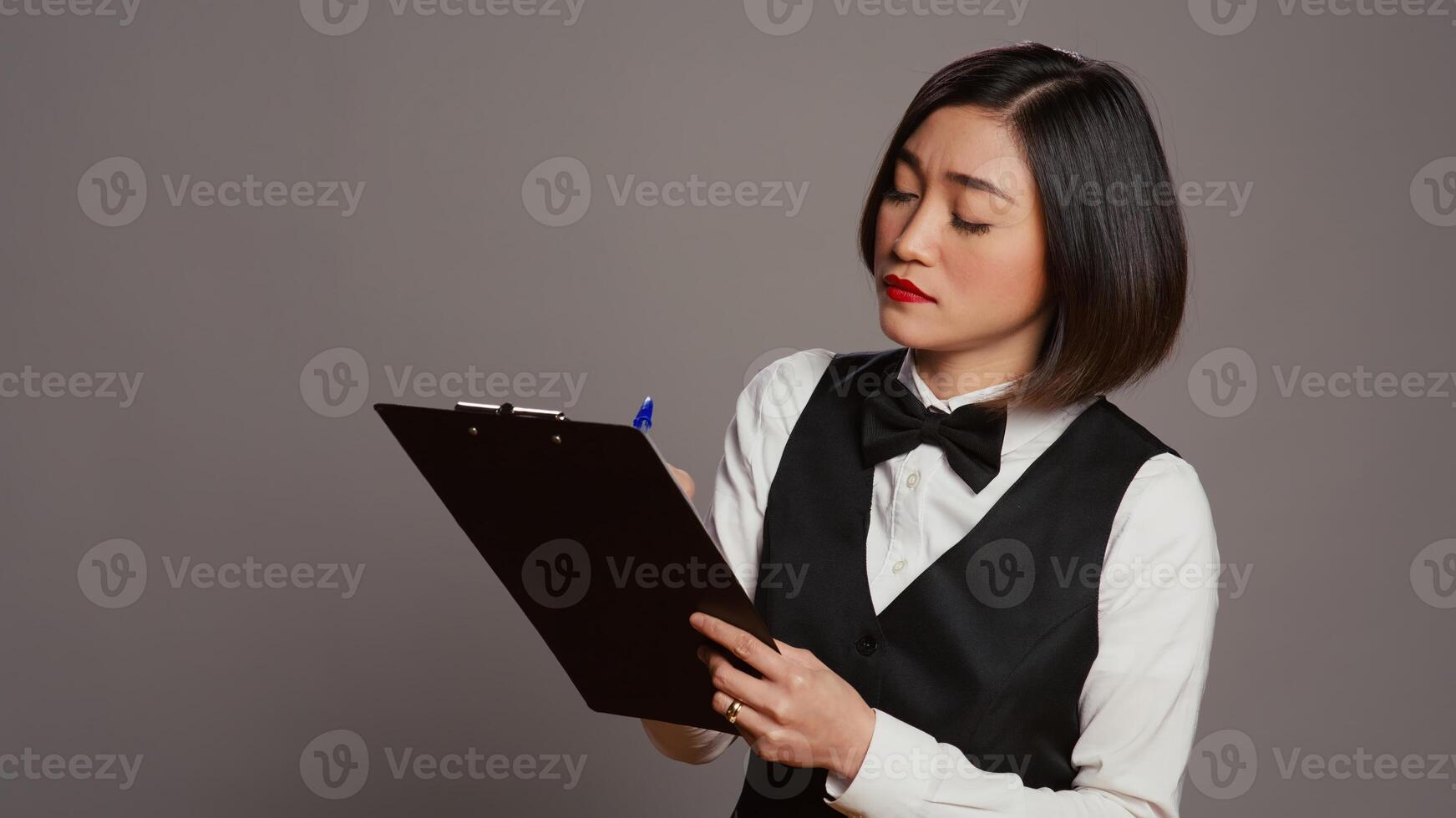 Hotel administrator checking off names of room reservations list, working in hospitality industry to greet clients. Asian receptionist examining registration notes on clipboard files. Camera B. photo