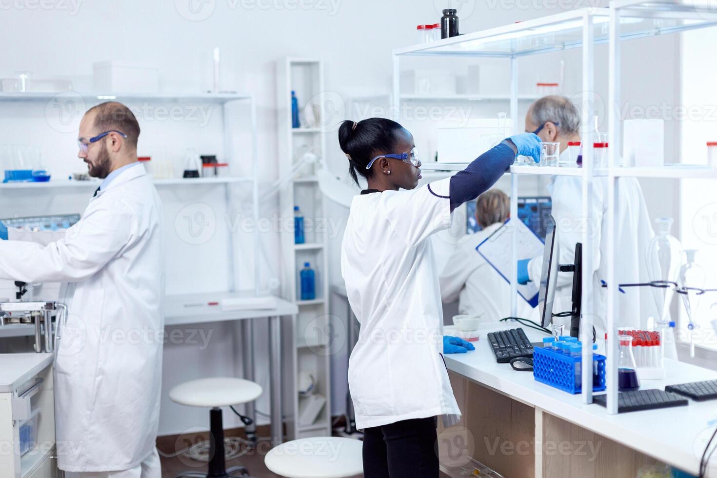African chemist in professional laboratory taking a glass flask with solution from shelf. Multiethnic team of researchers working in microbiology lab testing solution for medical purpose. photo