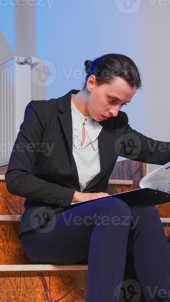 Overworked tired businesswoman reading financial raport of deadline project in the evening sitting on staircase of business building. Serious entrepreneur working on corporate job overtime late night photo