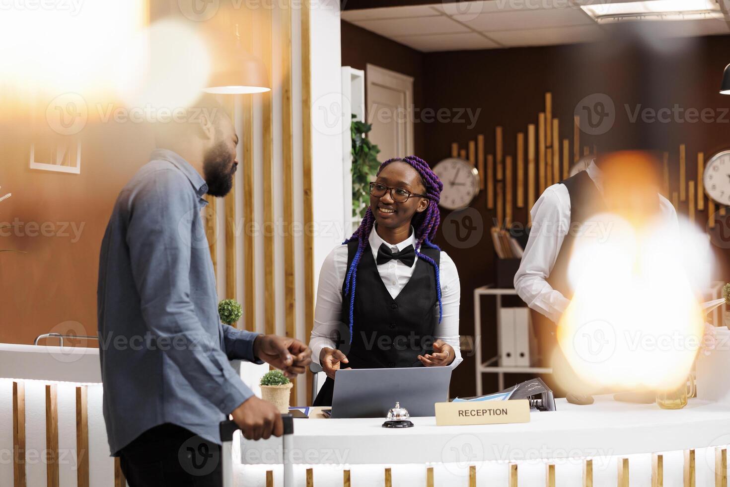 Smiling friendly African American woman receptionist talking with man guest at hotel reception. Black guy tourist standing with baggage at front desk doing check-in, receiving tourist information photo