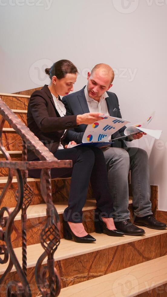 Overworked businessman reading raport about deadline financial crisis with coworker sitting on stairs. Colleague entrepreneur working together in evening at corporate job explaining difficult project. photo
