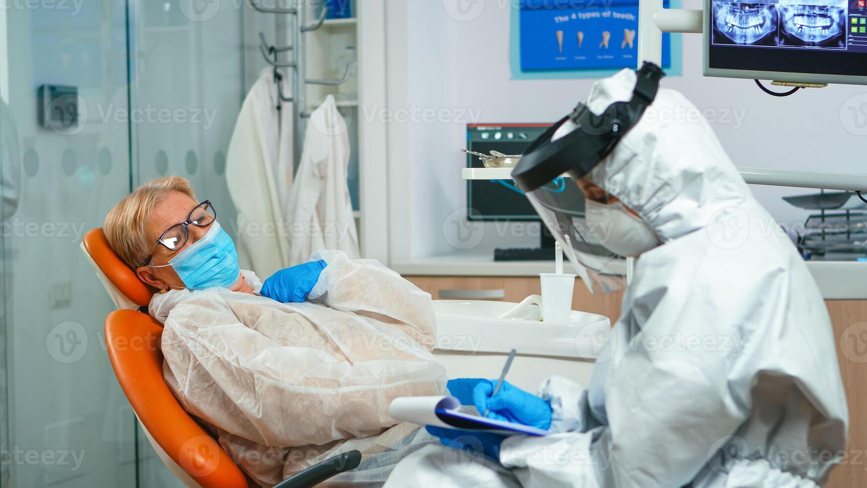 Dentist assistant in coverall interrogating senior woman during global coronavirus pandemic writing on clipboard. Concept of new normal orthodontic visit in coronavirus outbreak wearing protective suit and face shield photo