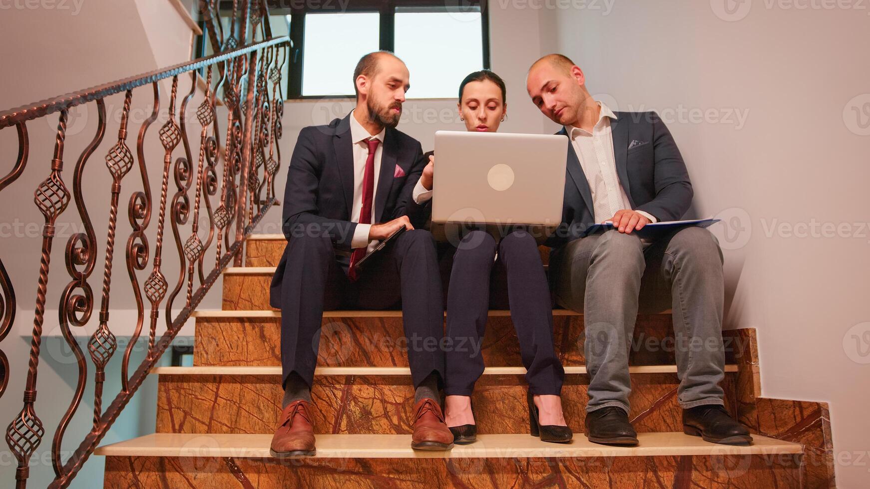 Overworked business colleagues doing overtime sitting on stairs using laptop and tablet, analysing difficult finance project. Company manager and office executive on stairs in corporate building. photo