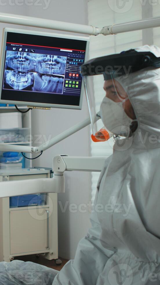 Dentist doctor with face shield pointing on stomatological monitor explaining dental x-ray to patient during global pandemic. Nurse and orthodontist wearing protection suit, coverall, mask and gloves photo
