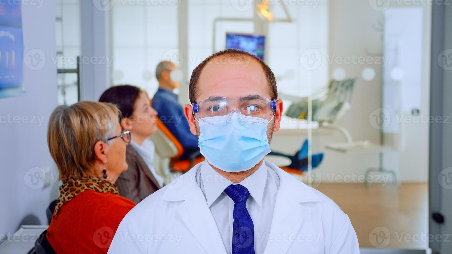 Portrait of tired stomatologist wearing protective mask at camera being in dental office while patients waiting him in background. Dentist looking on webcam sitting on chair in stomatological clinic. photo
