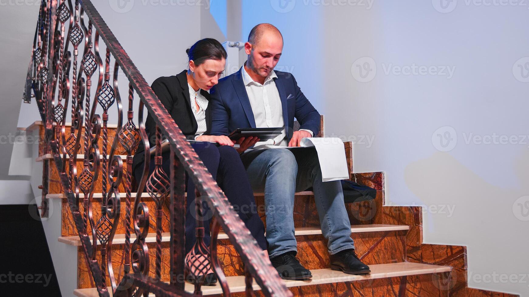 Business partners working with documents and tablet sitting on stairs doing overtime in corporate building. Colleagues entrepreneur studing late together at corporate job overworking. photo