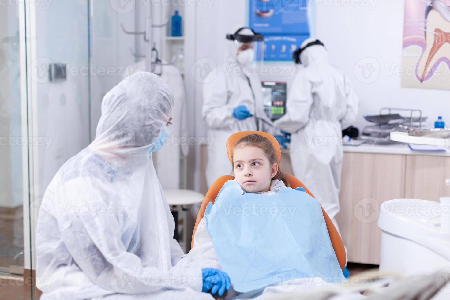 Little girl wearing coverall in dentist office waiting dental treatment sitting on chair. Stomatologist during covid19 wearing ppe suit doing teeth procedure of child sitting on chair. photo