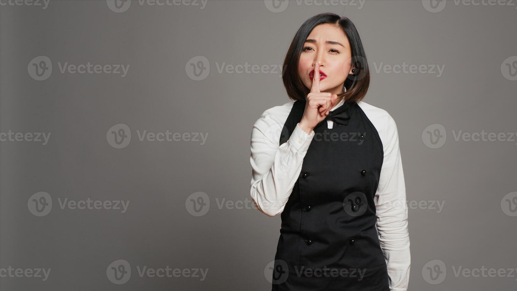 Restaurant hostess showing silence symbol in studio, doing hush mute gesture with finger over lips. Private waitress asking to keep secret with confidential information, secrecy sign. Camera A. photo