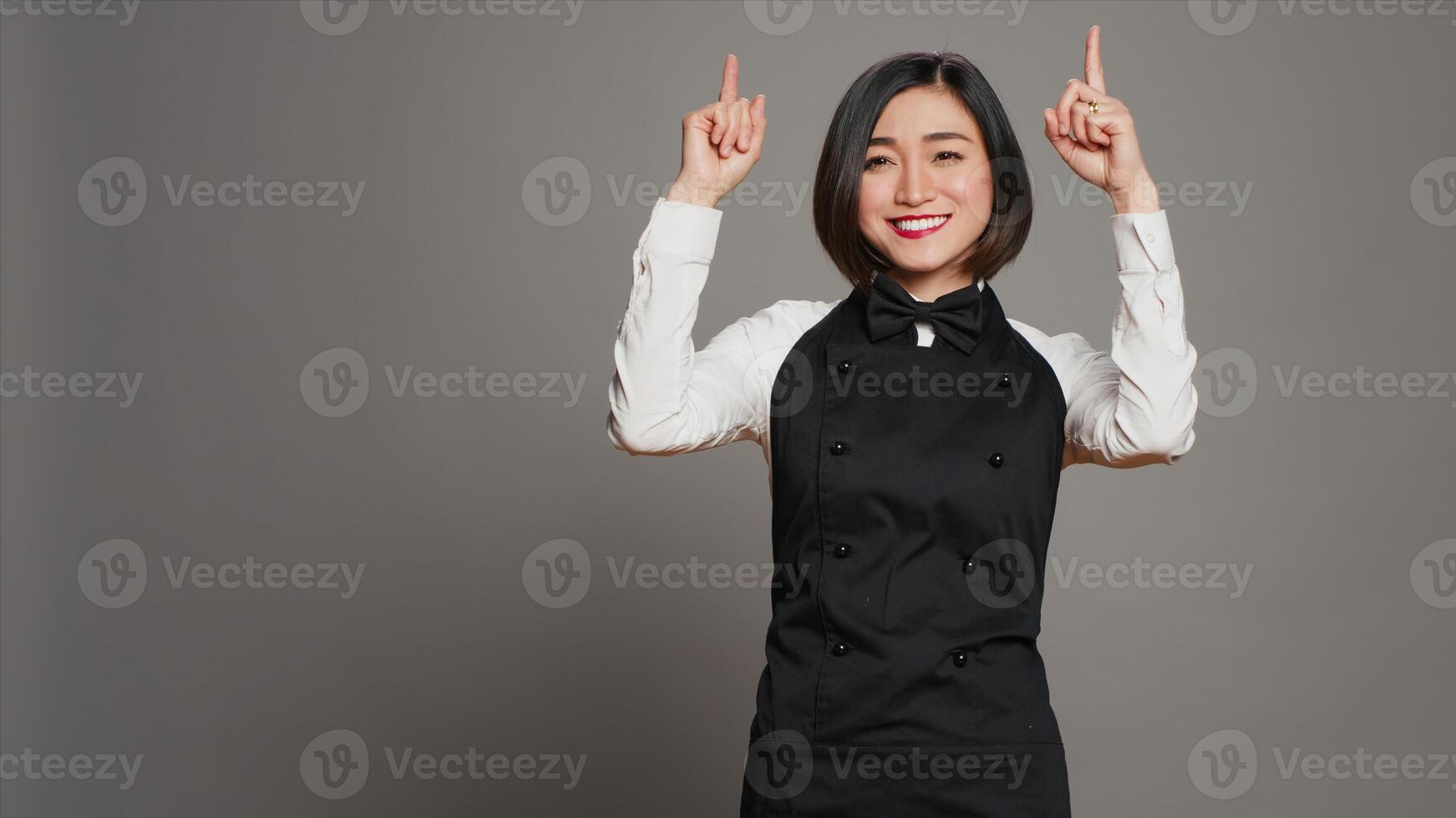 Asian woman pointing at something upwards in studio, indicating direction above her head. Waitress server with apron showing up indication over grey background, table service. Camera A. photo