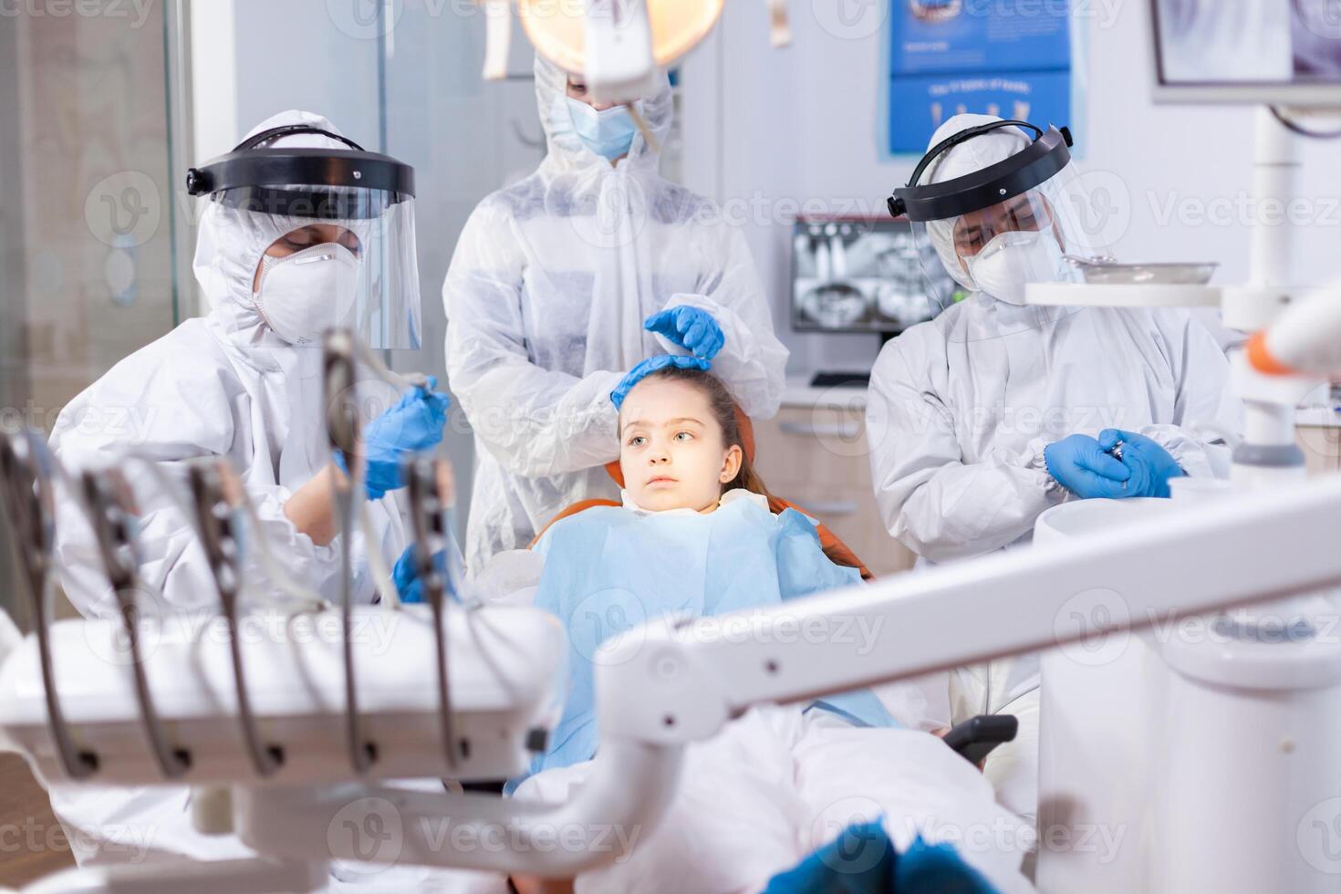 Mother dressed in ppe suit comforting daughter in the course of dental treatment. Stomatology team wearing ppe suit during covid19 doing procedure on child teeth. photo
