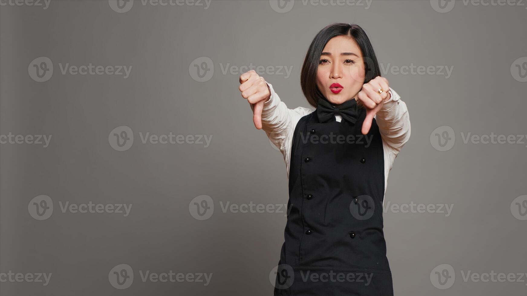 Asian restaurant hostess showing thumbs down and dislike sign, expressing negativity and disapproval in studio. Displeased waitress working at a diner and serving customers. Camera A. photo