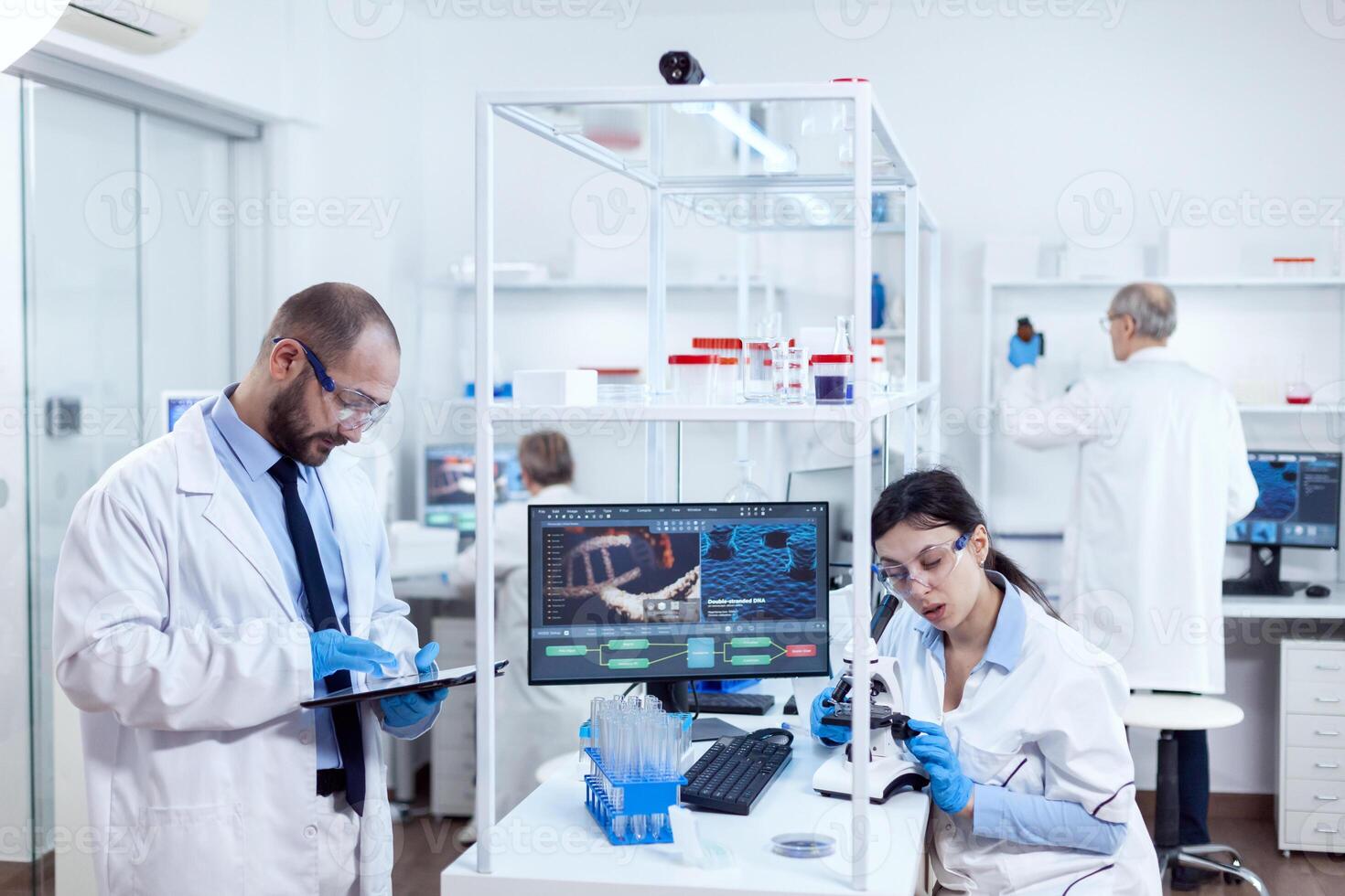 Scientist analysing test results on tablet pc. Team of researchers doing pharmacology engineering in sterile laboratory for healthcare industry with african assistant in the background. photo