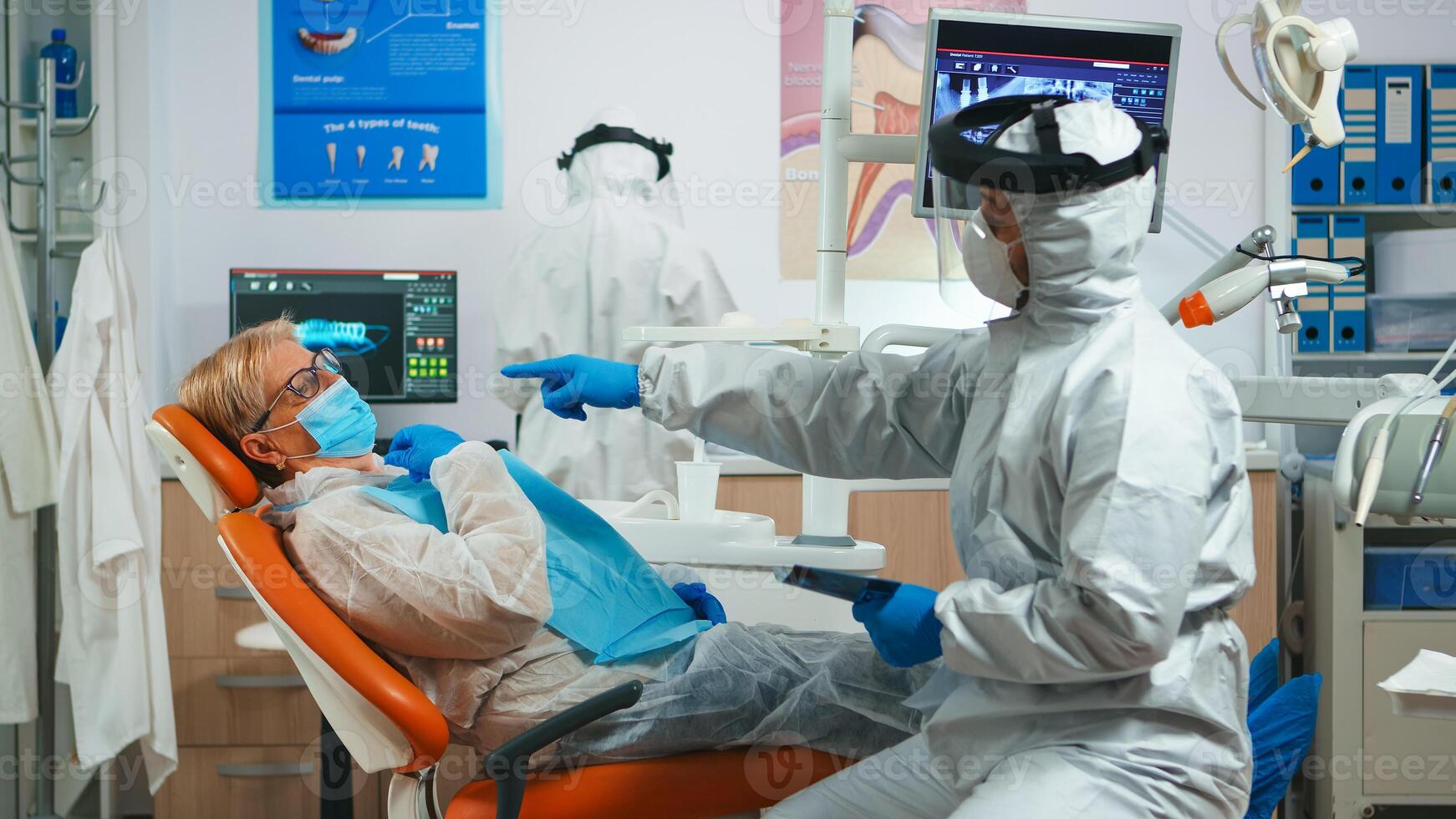 Dentistry doctor in protection equipment explaining dental problem using x-ray during covid-19. Assistant and dentist talking with senior patient wearing suit, coverall, face shield, mask and gloves. photo