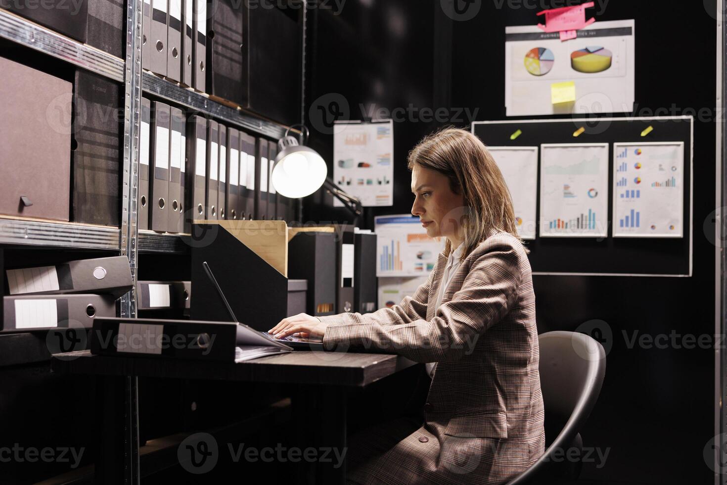 Woman manager typing administration report on laptop computer, working overtime at management research in file room. Bookkeeper checking accountancy record, searching for business documentation photo