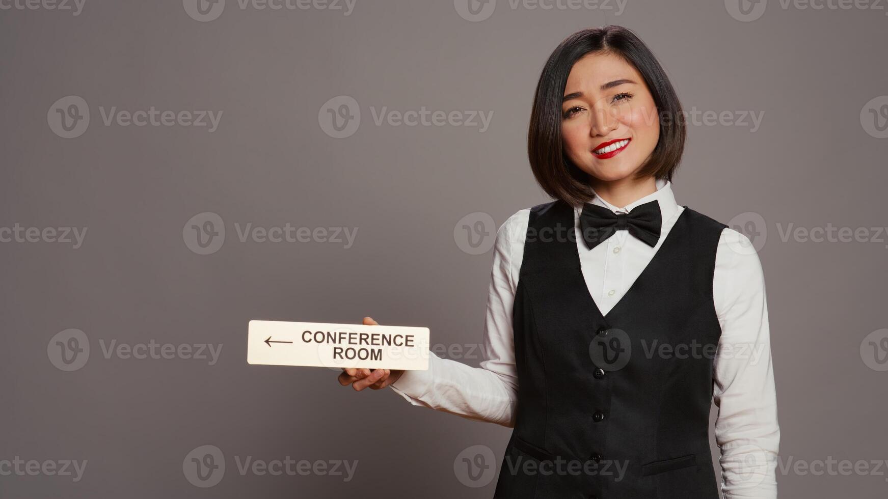 Asian receptionist pointing at wall indicator for conference room, using pointer to help customers find all amenities and facilities. Woman employee holding sign to indicate directions. Camera B. photo