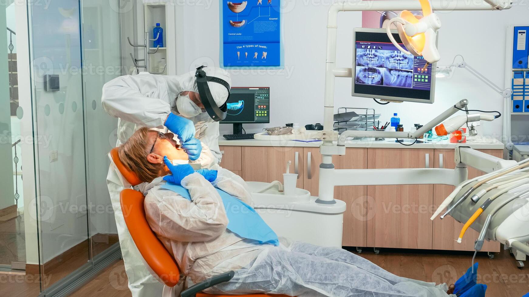 Dentist doctor in coverall finishing dental treatment in stomatological clinic during global pandemic. Assistant and orthodontic wearing protection suit, face shield mask and gloves examining patient photo
