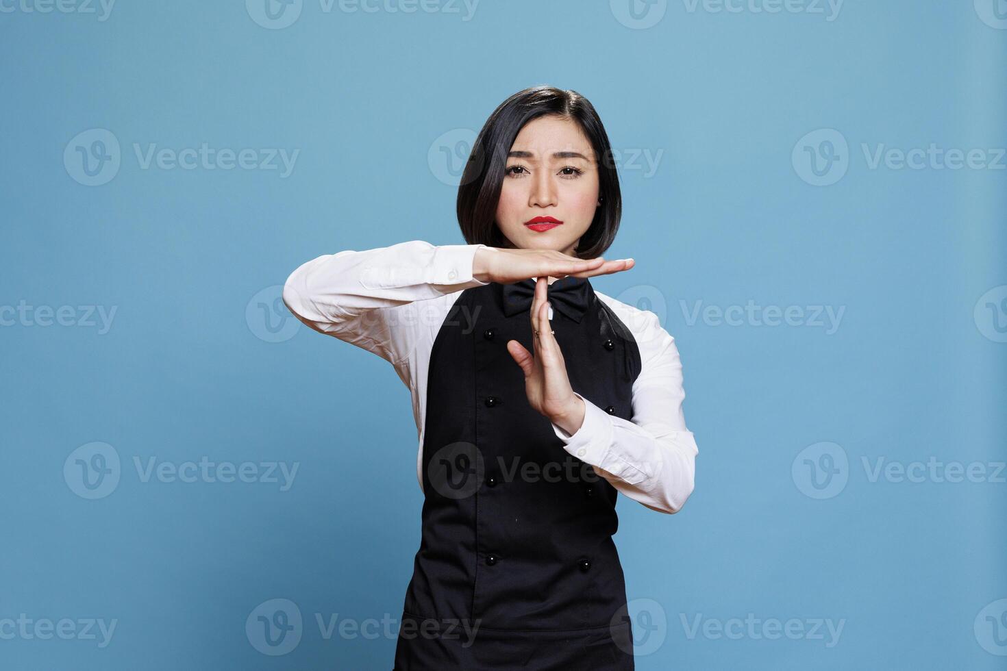 Confident asian waitress dressed in restaurant uniform making timeout gesture with arms portrait. Woman receptionist showing take a break sign with hands and looking at camera with serious expression photo