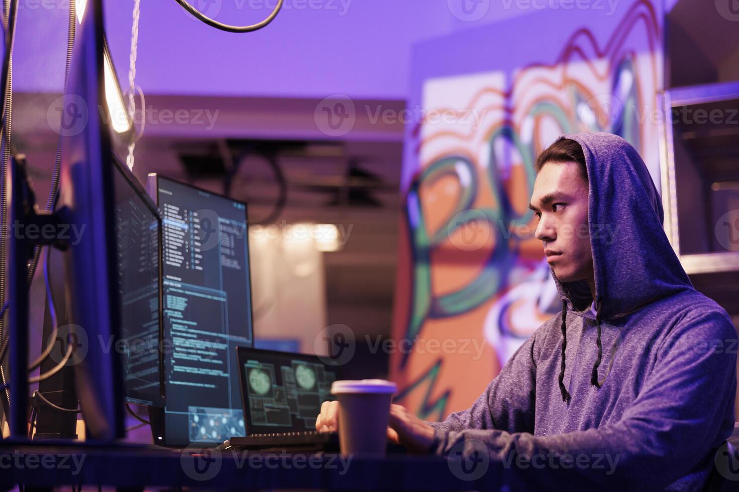 Asian hacker stealing information from server using computer worm. Criminal in hood coding illegal internet virus and breaking into server while drinking coffee to go in dark room photo