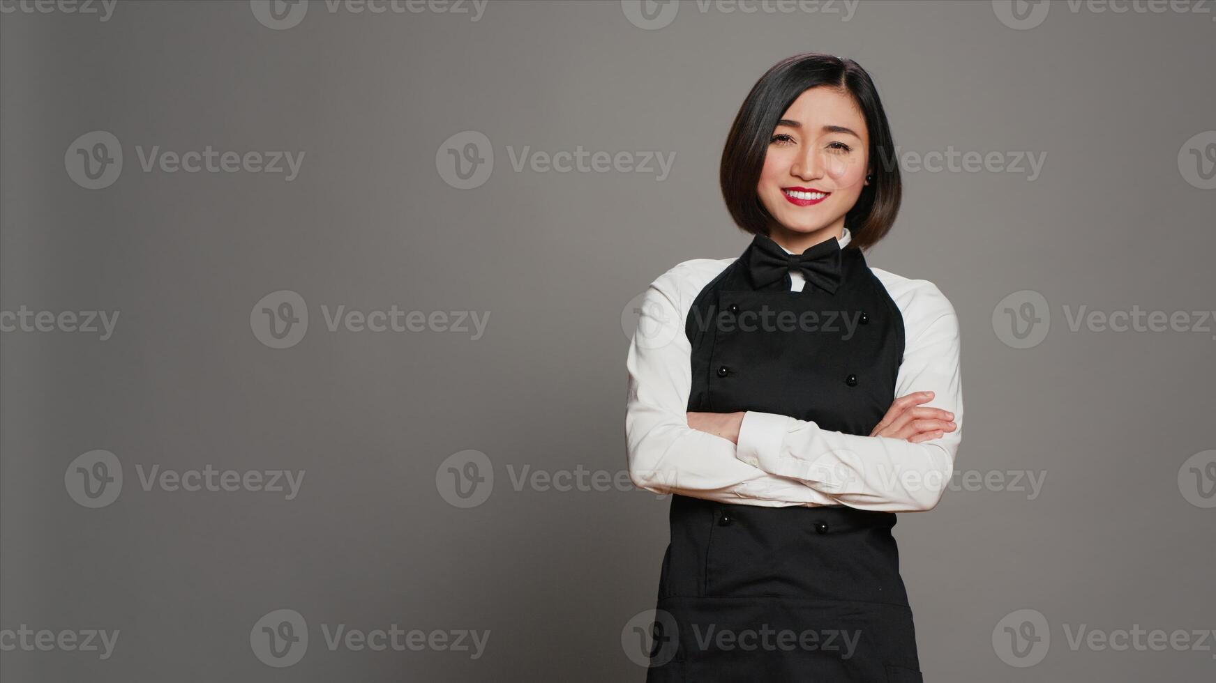 Restaurant hostess posing with confidence in studio, standing with arms crossed over grey background. Asian waitress barista with gourmet serving expertise smiling on camera. Camera A. photo