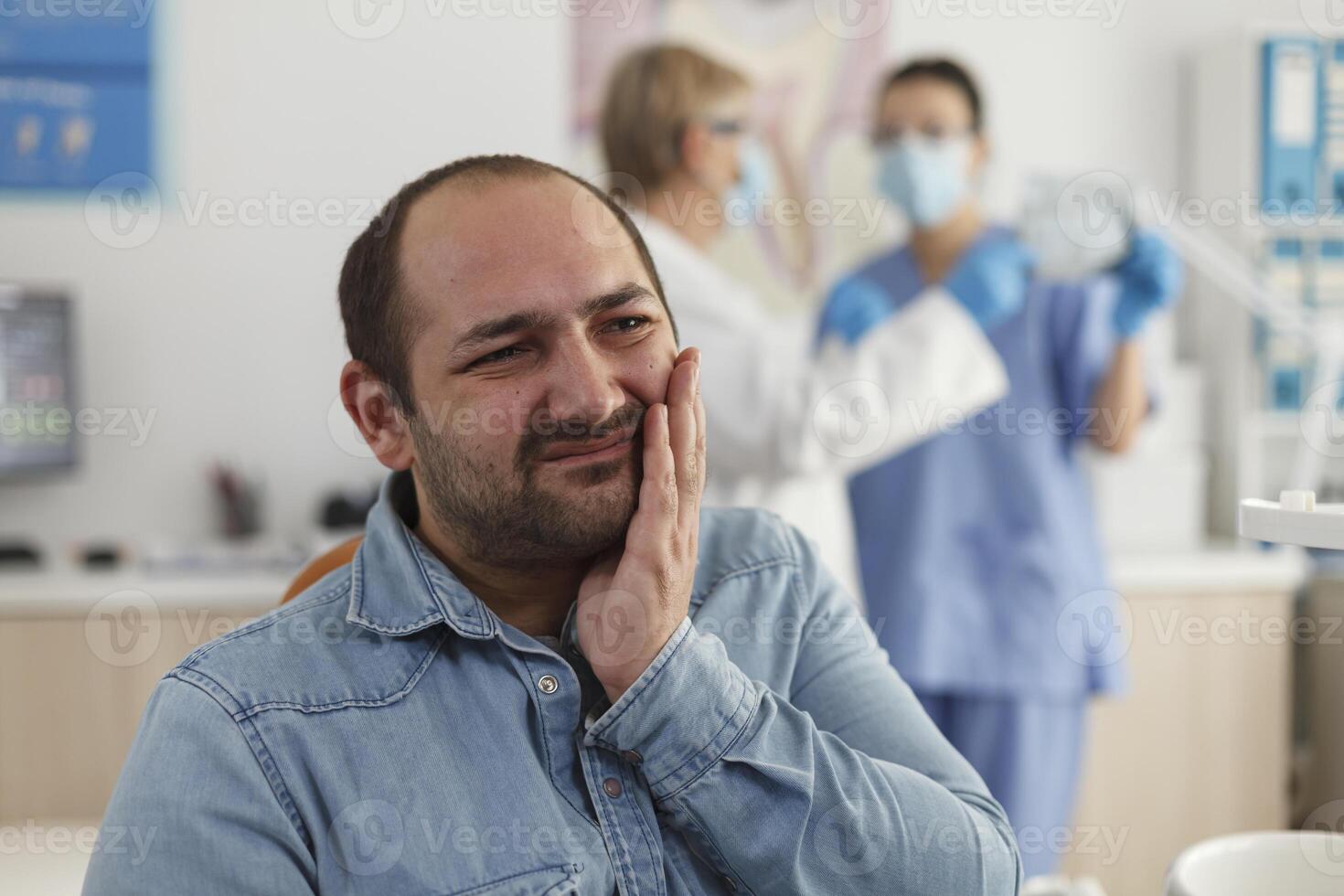 Close up of man patient with toothache sitting on dental chair during stomatological examination waiting for medical expertise. In background specialist team discussing dentistry treatment photo