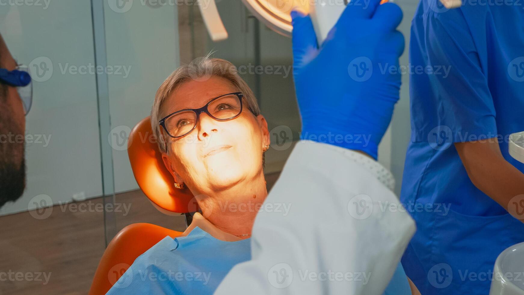 Patient on the chair in dentist surgery treating with a dental drill. Orthodontist lighting the lamp and speaking to patient sitting on stomatological chair while nurse preparing tools for surgery. photo