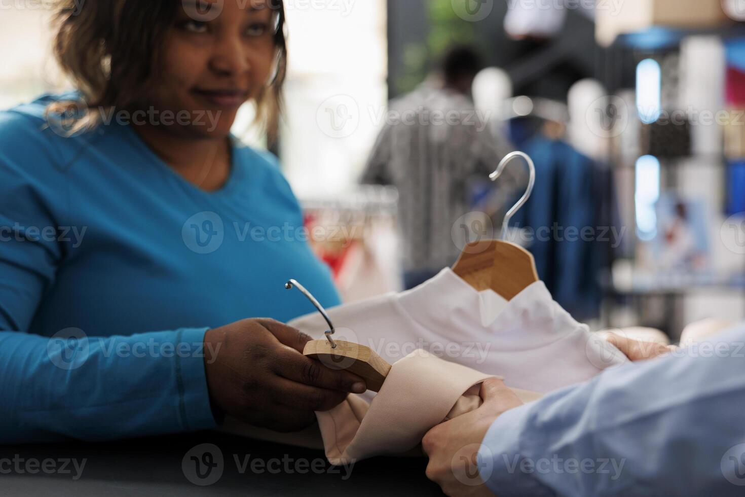 African american woman shopping for casual wear, holding multiple shirts in modern boutique. Stylish customer standing at counter desk, discussing items fabric with store employee. Fashion concept photo