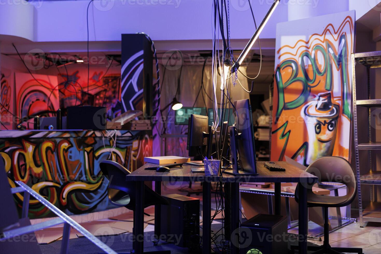 Internet criminals abandoned warehouse with graffiti at night time. Computers on desks and equipment for hacking and malware coding in hackers dark hideout room with neon light photo