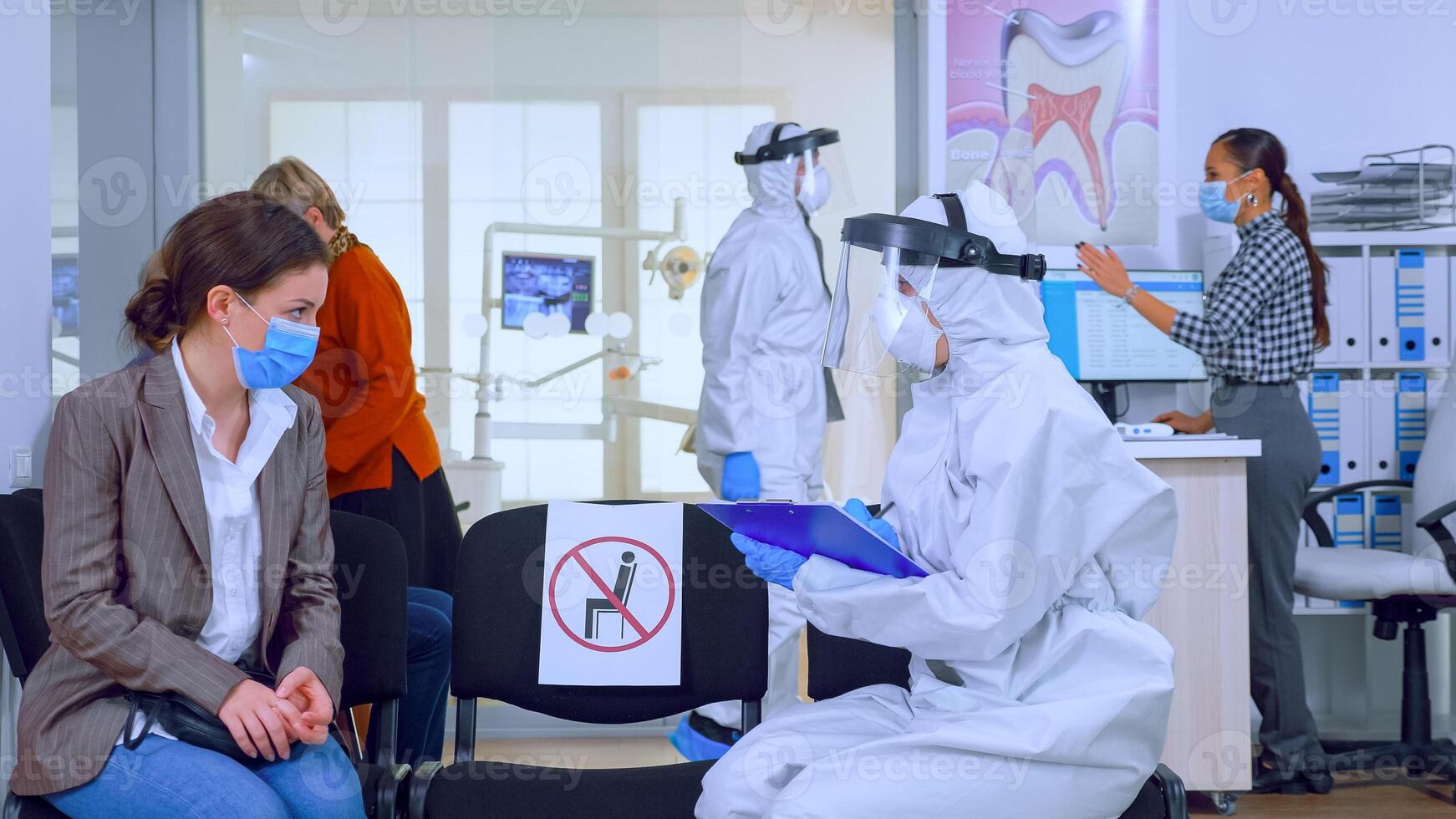Dentist assistant with ppe equipment talking with patient before consultation during coronavirus epidemic sitting on chairs in waiting area keeping distance. Concept of new normal dentist visit. photo