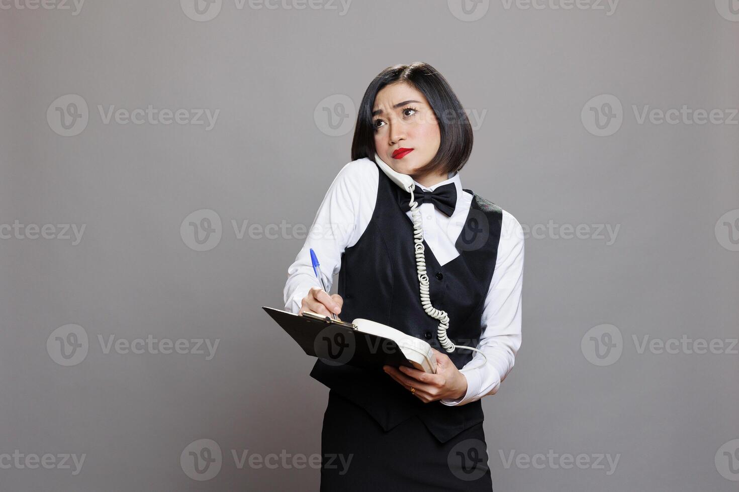 Catering service employee frowning while speaking on landline phone with client and taking notes on clipboard. Waitress looking upwards while chatting with cafe manager on telephone photo