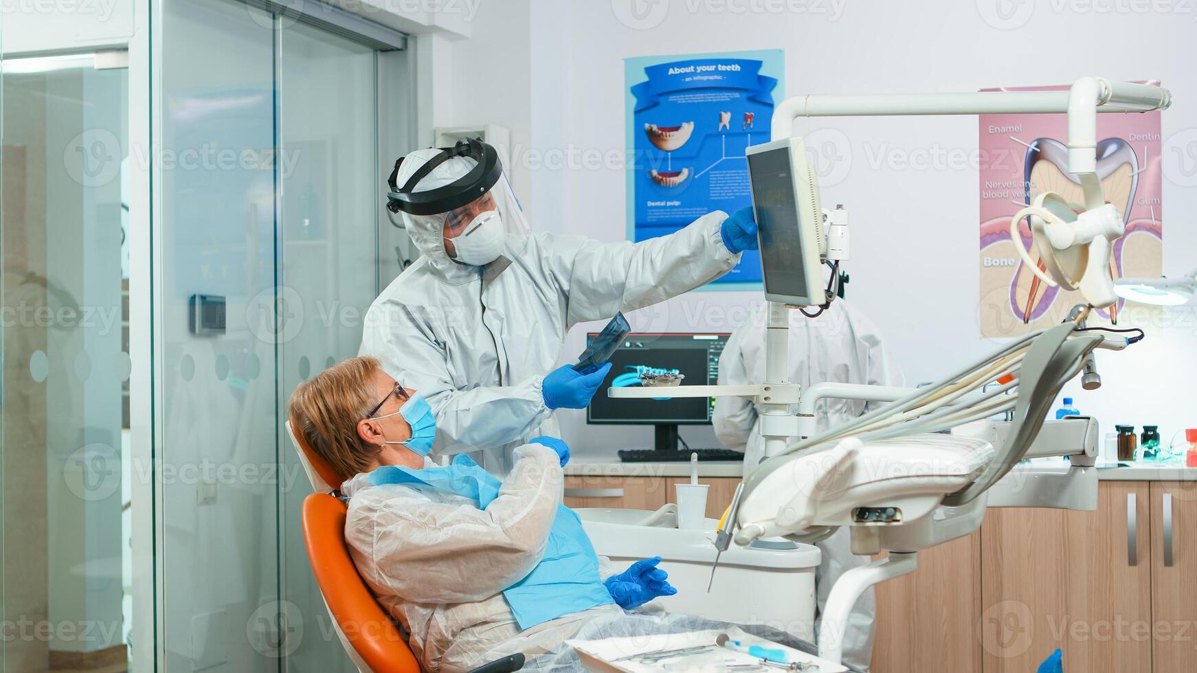 Dentist in coverall showing patient situation of teeth x-ray image during global pandemic. Assistant and doctor talking with senior woman wearing protection suit, face shield, mask and gloves. photo
