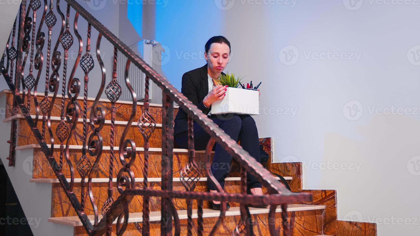 Frustrated business woman getting fired sitting on office staircase and crying holding box of stuff. Employee getting up on stair hugging her colleague, working in modern financial building. photo