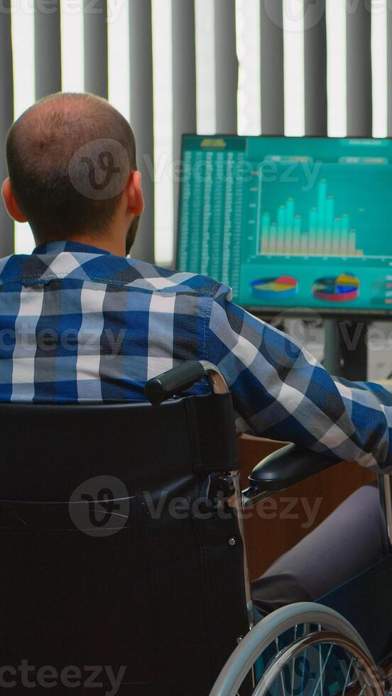 Disabled entrepreneur sitting in wheelchair immobilized analysing financial economy statistics on computer in business office discussing with colleague. Handicapped businessman using modern technology photo