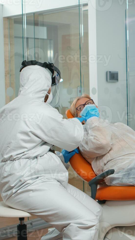 Dental specialist with coverall talking with senior patient before examination during pandemic covid-19. Concept of new normal dentist visit in coronavirus outbreak wearing protective suit and face shield photo