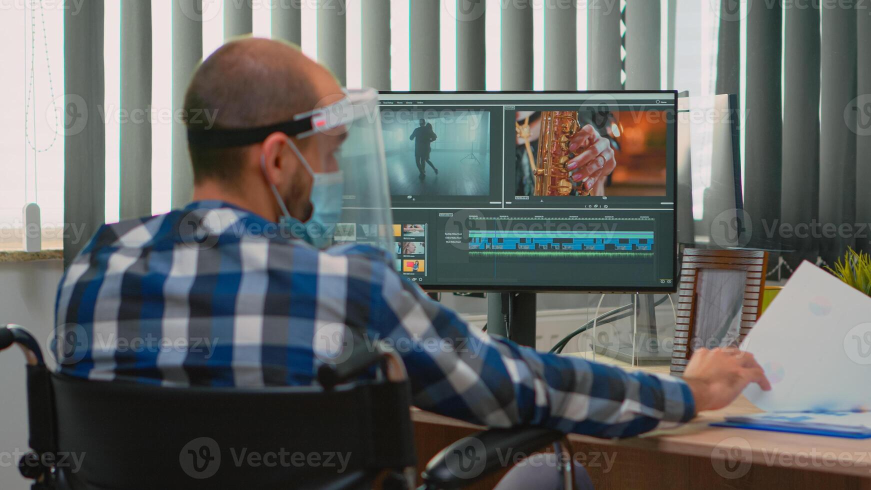 Disabled man videographer with face protection editing video project creating content sitting in wheelchair in new normal office. Blogger working durig global pandemic respecting social distance. photo