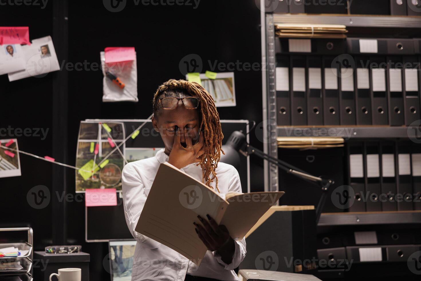 Tired woman detective rubbing eyes and working late in investigation agency office room. African american exhausted police investigator reading crime case file at night time photo
