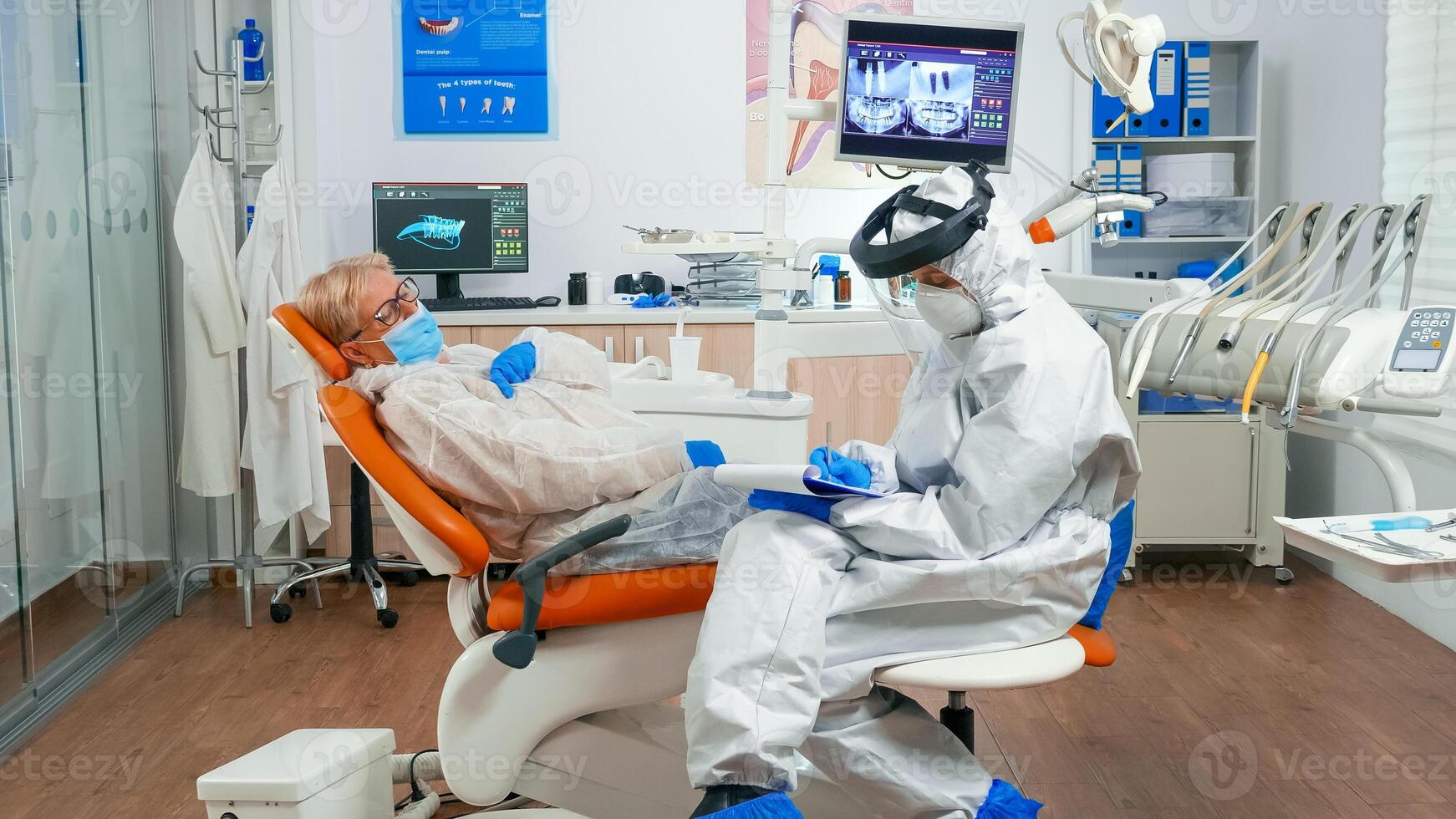 Dentist assistant in coverall talking with senior woman during global coronavirus pandemic writing on clipboard. Nurse and orthodontic doctor wearing face shield, protection suit, mask and gloves. photo